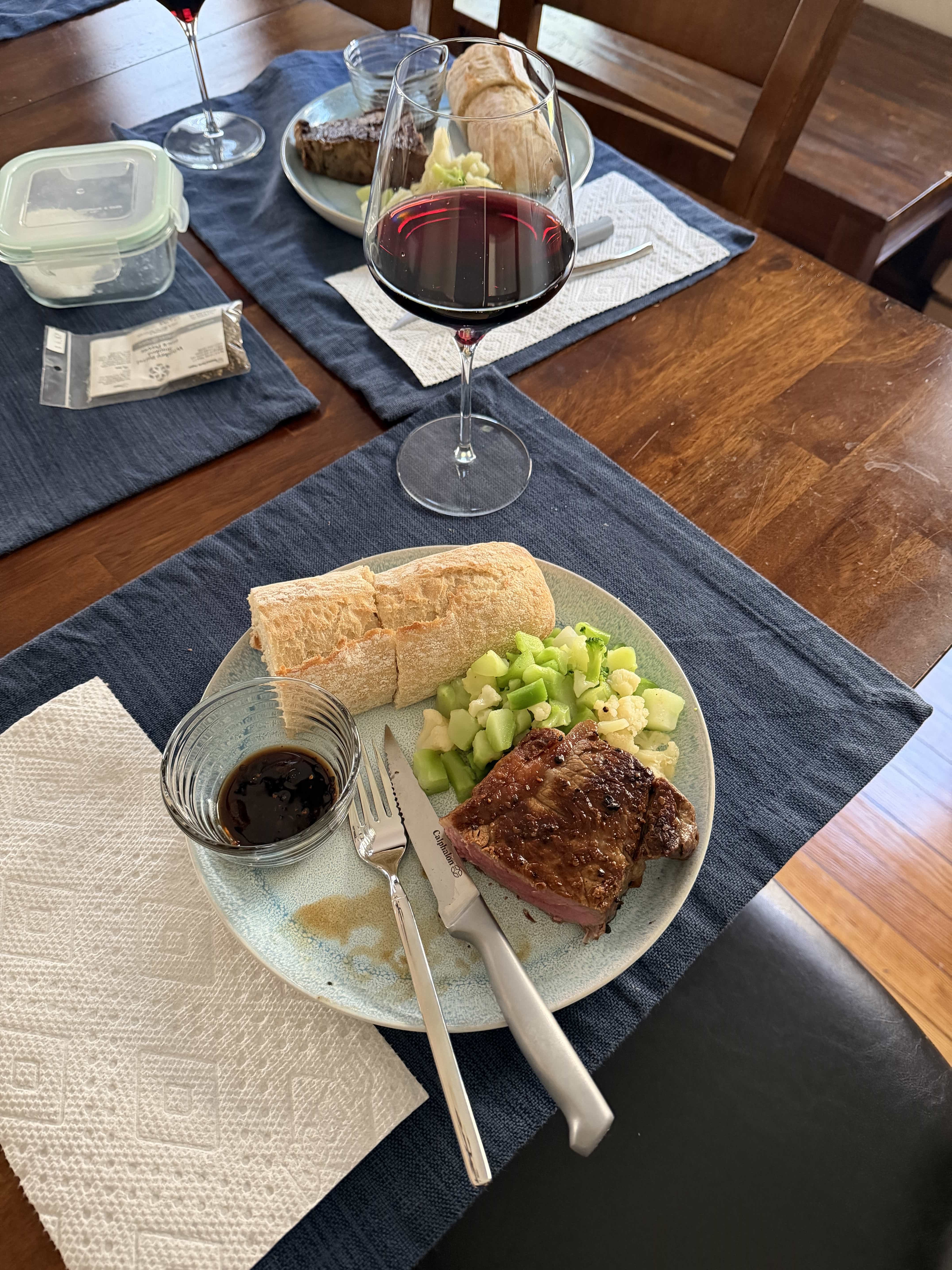 A dinner plate containing a cut steak, chopped broccoli and cauliflower, and part of a baguette. There is a glass of red wine and a similar plate in the background